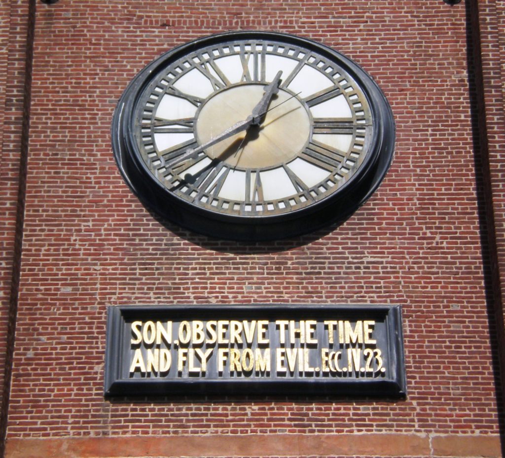 A white and brass clock face with black roman numerals on a brick wall. A black rectangle below the clock contains the inscription, in golden letters: Son, observe the time and fly from evil. Ecc.IV.23