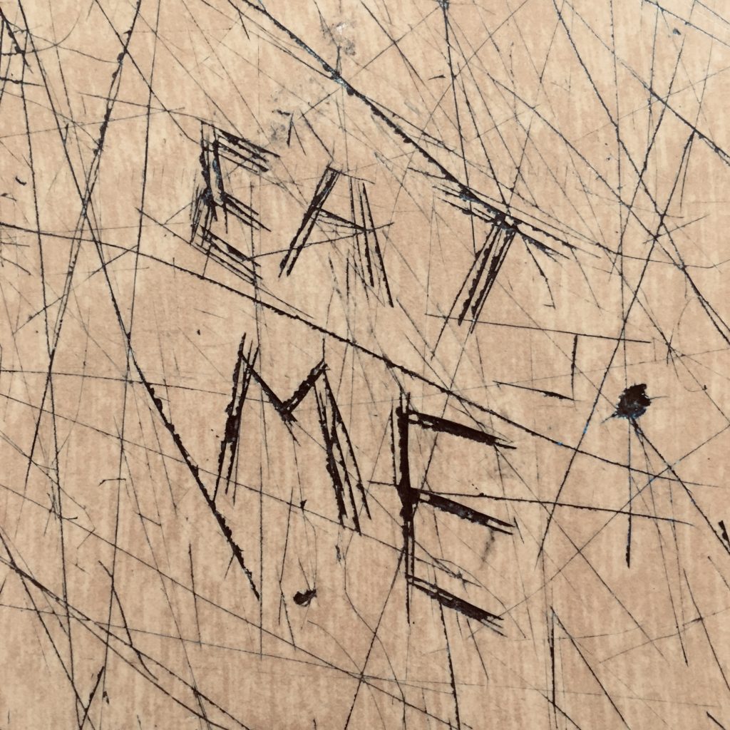 The words "Eat me" in all caps, scratched into the wood veneer of a desktop.